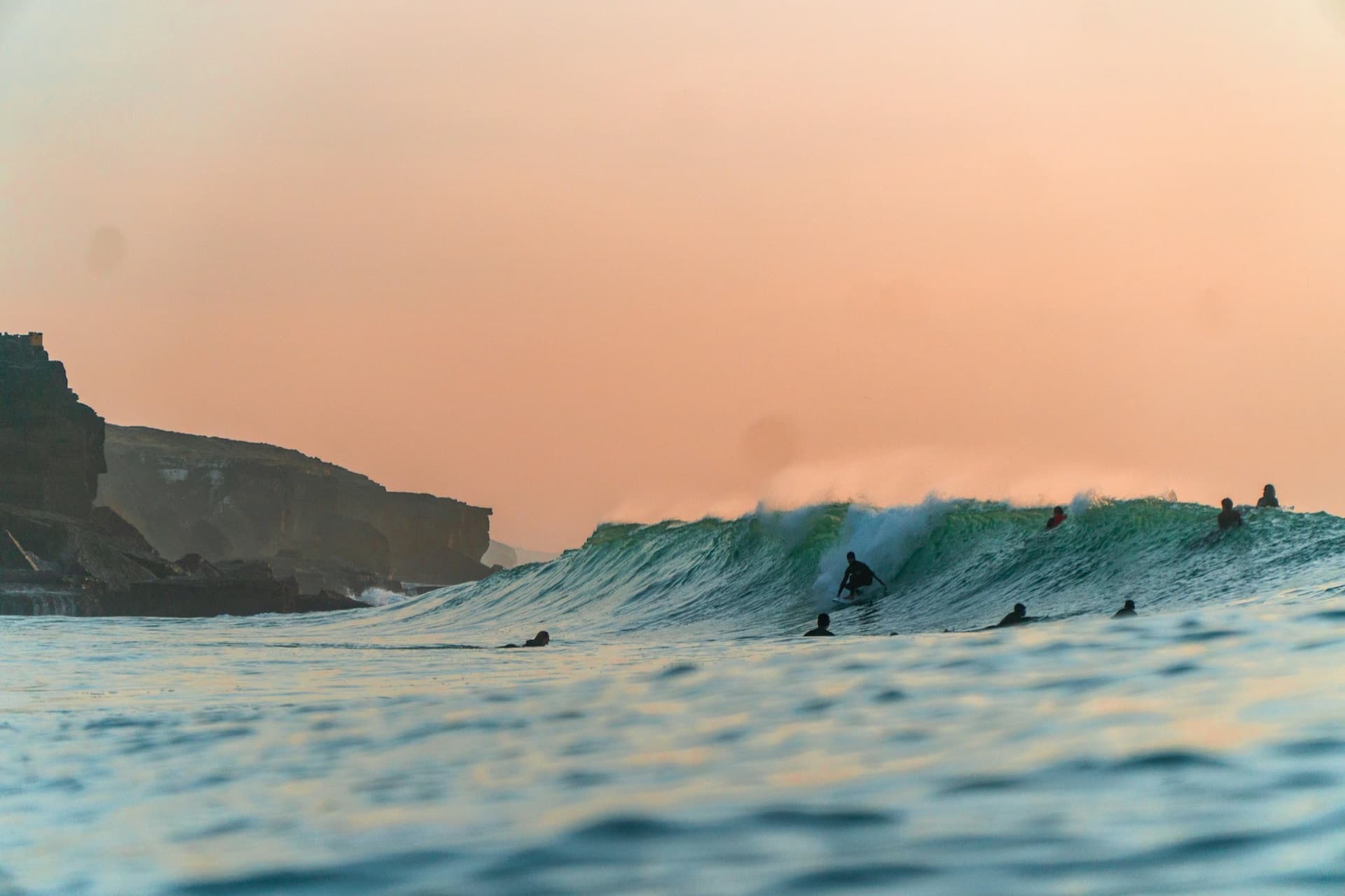 A surfer riding a peeling wave at Ericeira, Portugal, with cliffs behind under a pink sunset sky