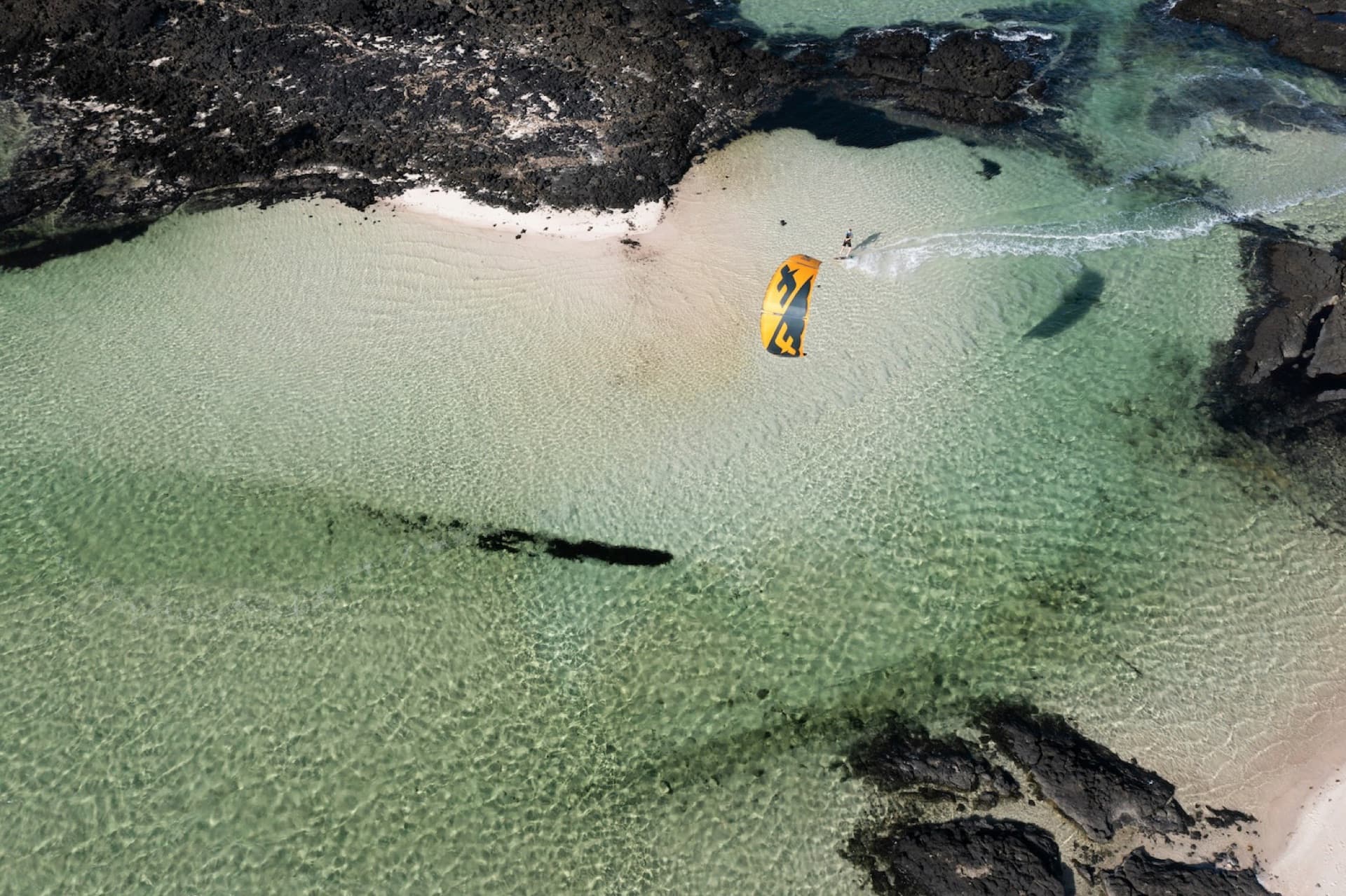 An aerial view of a kitesurfer crossing a shallow turquoise lagoon between black volcanic rocks on Fuerteventura
