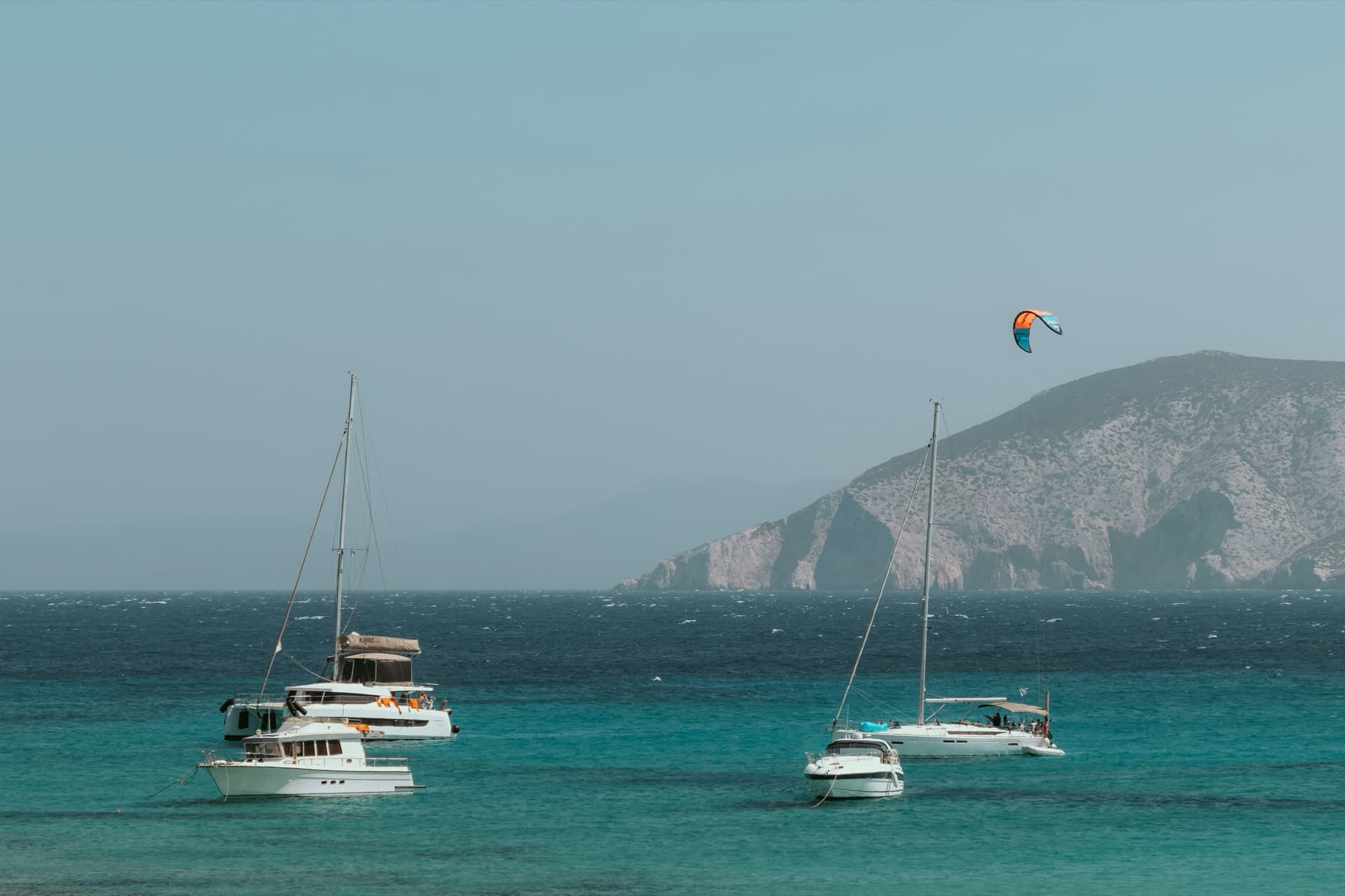 Boats anchored in turquoise Aegean water with a kite up off a rocky Cycladic headland