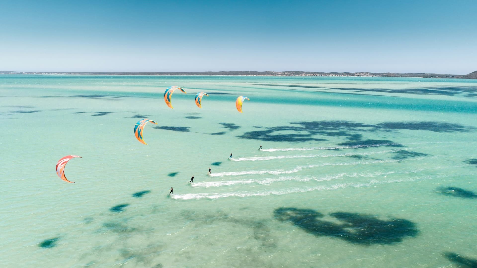 Several kitesurfers riding across a bright turquoise shallow lagoon, kites of different sizes in the sky