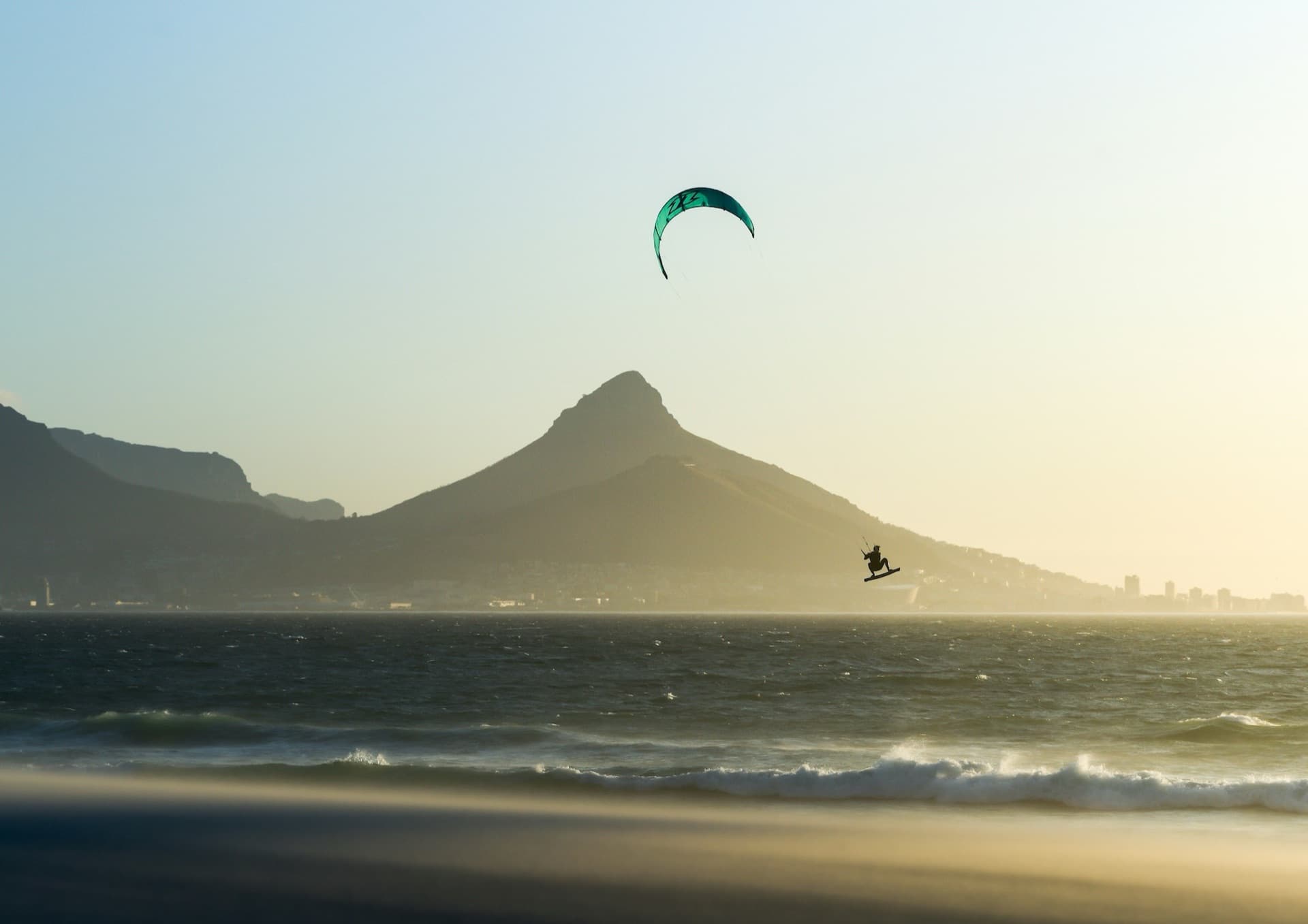 A kitesurfer boosting a jump off the coast at golden hour, kite high overhead, a mountain behind