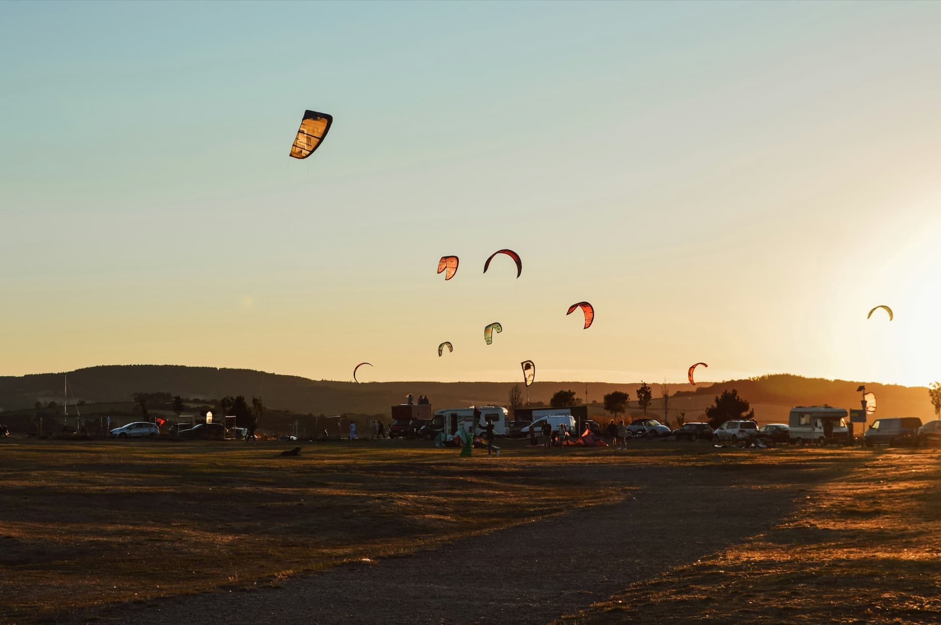 Kitesurfing kites in the air over a launch area at sunset, vans parked on the grass and low hills on the horizon