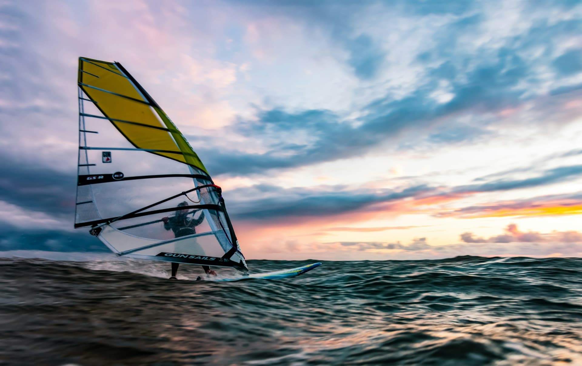 A windsurfer planing across choppy blue water on a sunny day, sail powered up and board on the rail